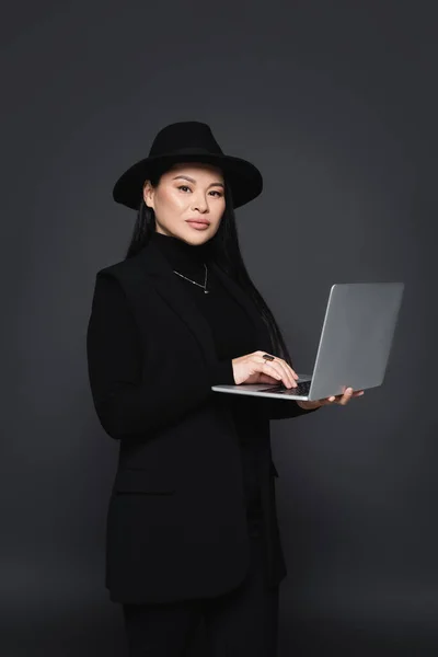 Moda mujer asiática en sombrero fedora y chaqueta usando portátil aislado en gris oscuro - foto de stock