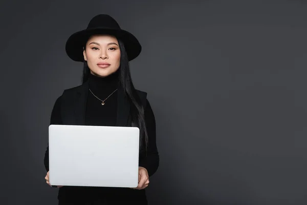 Stilvolle asiatische Frau mit Fedora-Hut und Rollkragen hält Laptop isoliert auf dunkelgrau — Stockfoto