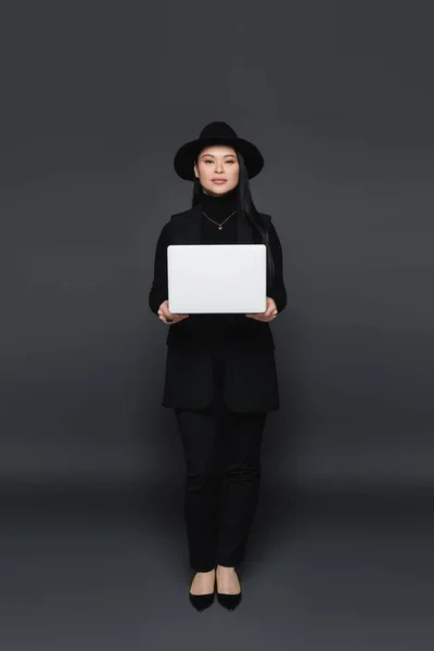 Full length of stylish asian woman in fedora hat holding laptop on dark grey background — Stock Photo