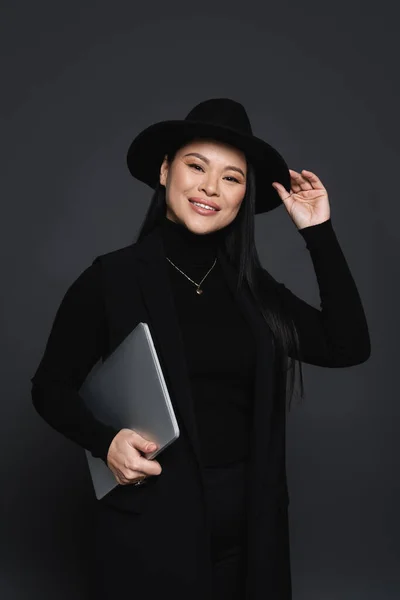 Alegre ásia mulher no fedora chapéu e gola alta segurando laptop isolado no escuro cinza — Fotografia de Stock