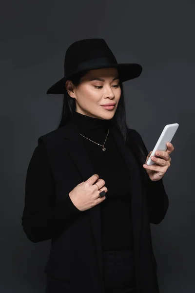 Femme asiatique élégante en chapeau fedora en utilisant un smartphone isolé sur gris foncé — Photo de stock