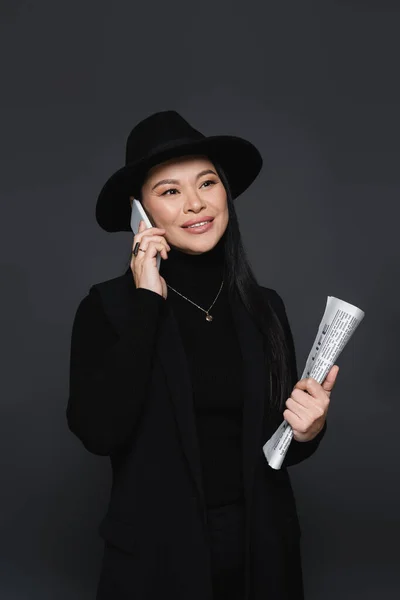 Elegante mujer asiática hablando en smartphone y sosteniendo periódico aislado en gris oscuro - foto de stock