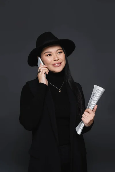 Stylish asian woman talking on smartphone and holding newspaper isolated on dark grey — Stock Photo