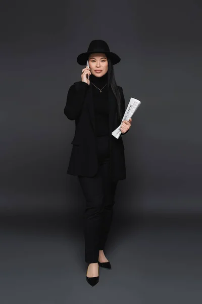 Volle Länge von trendigen asiatischen Frau spricht auf Smartphone und hält Zeitung auf dunkelgrauem Hintergrund — Stockfoto