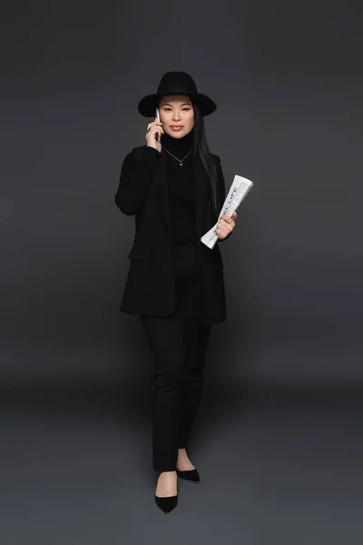 Full length of trendy asian woman talking on smartphone and holding newspaper on dark grey background — Stock Photo