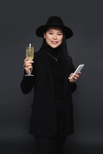 Positivo elegante asiático mulher segurando smartphone e copo de champanhe isolado no cinza escuro — Fotografia de Stock