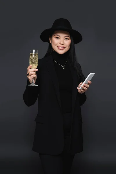 Positive stylish asian woman holding smartphone and glass of champagne isolated on dark grey — Stock Photo