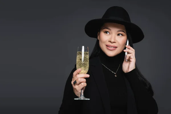 Elegant asian woman in fedora hat talking on smartphone and holding champagne isolated on dark grey — Stock Photo