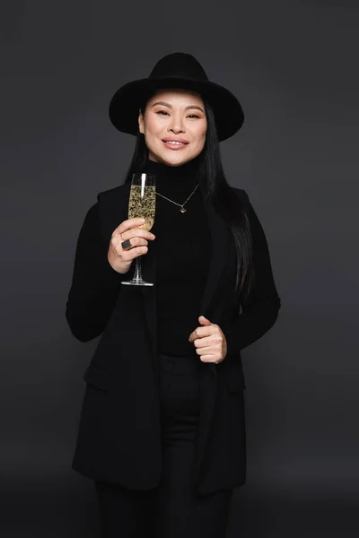 Positive asian woman in fedora hat holding champagne isolated on dark grey — Stock Photo