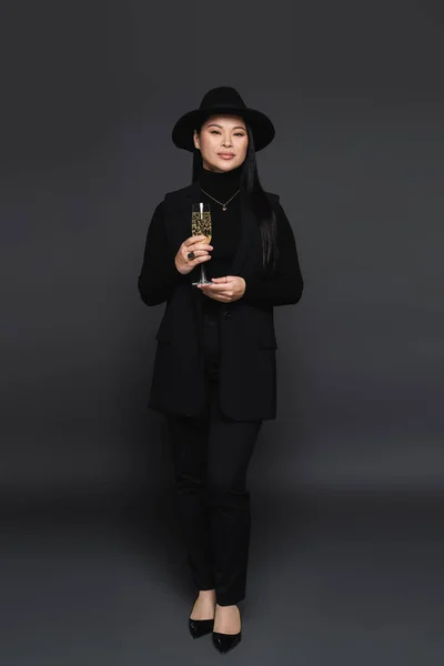 Comprimento total de elegante modelo asiático em chapéu fedora segurando champanhe no fundo cinza escuro — Fotografia de Stock