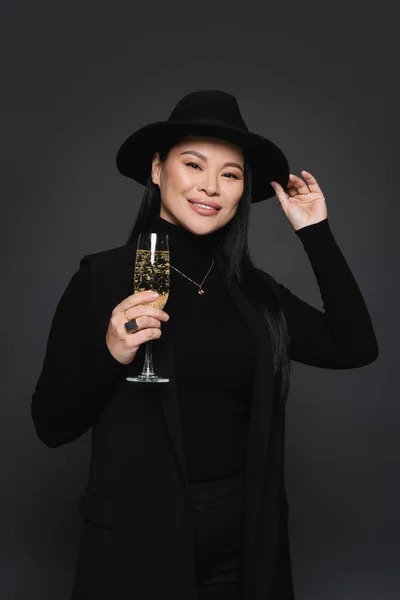 Smiling asian woman in fedora hat and blazer holding glass of champagne isolated on dark grey — Stock Photo