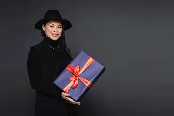 Smiling asian woman in fedora hat holding present isolated on dark grey — Stock Photo