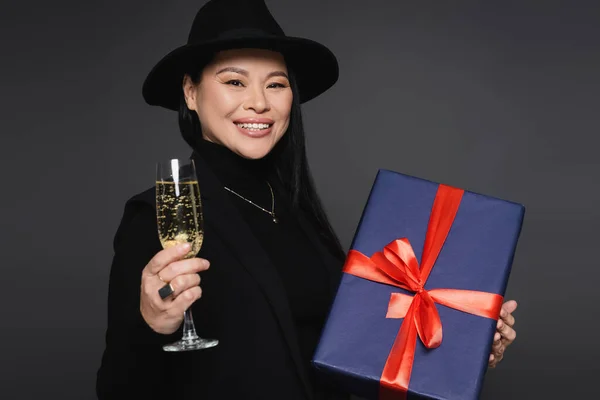 Cheerful asian woman in blazer and fedora hat holding champagne and present isolated on dark grey — Stock Photo