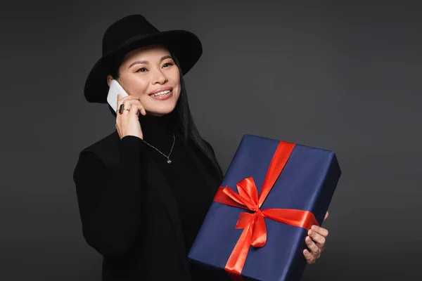 Позитивна азіатська жінка в капелюсі Фургори говорить на смартфоні і тримає подарунок ізольовано на темно-сірому — стокове фото