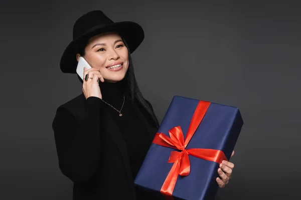 Positivo asiatica donna in cappello fedora parlando su smartphone e tenendo il regalo isolato su grigio scuro — Foto stock