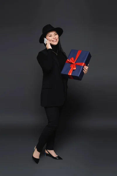 Полная длина модная азиатская женщина говорит на смартфоне и держит подарочную коробку на темно-сером фоне — стоковое фото