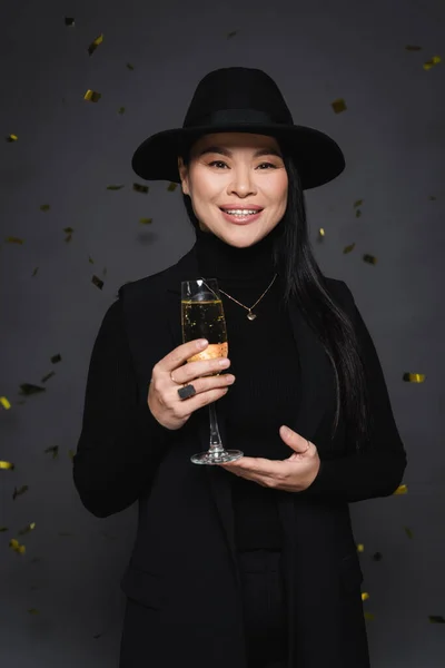 Felice donna asiatica in cappello fedora che tiene champagne vicino coriandoli su sfondo grigio scuro — Foto stock