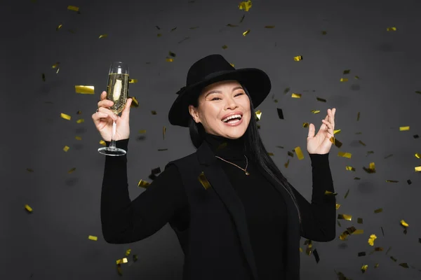 Positive asian woman in fedora hat holding champagne under falling confetti on dark grey background — Stock Photo
