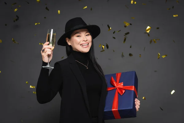 Positivo asiático mujer en fedora sombrero celebración champán y presente bajo confeti sobre oscuro gris fondo - foto de stock
