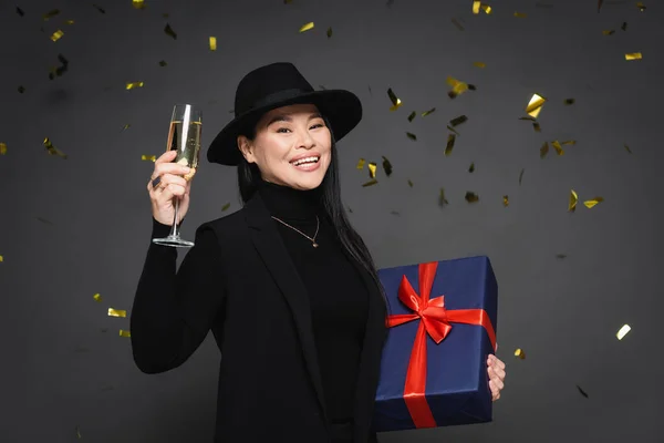 Positive asian woman in fedora hat holding champagne and present under confetti on dark grey background — Stock Photo