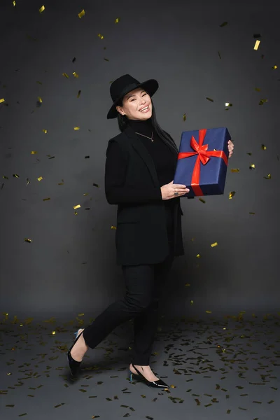 Pleine longueur de positif asiatique femme en fedora chapeau tenant cadeau sous tomber confettis sur fond gris foncé — Photo de stock