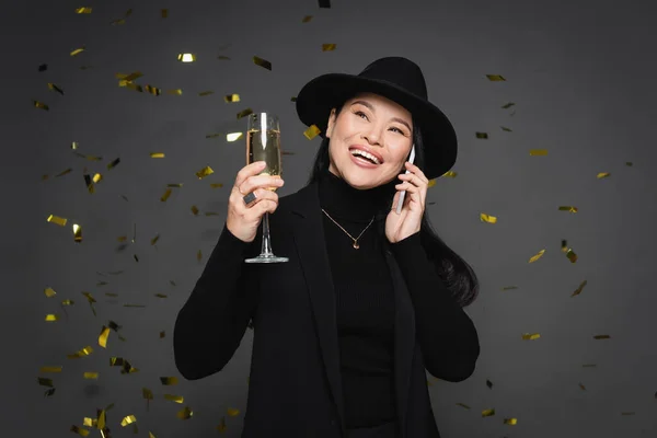 Joyful asian woman in fedora hat talking on smartphone and holding champagne under falling confetti on dark grey background — Stock Photo