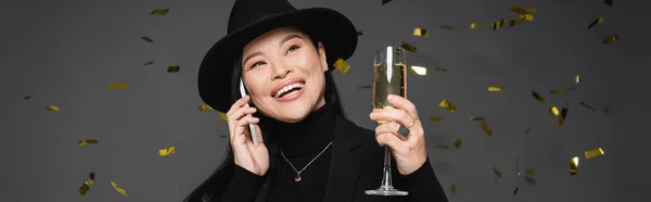 Щаслива азіатська жінка в капелюсі Фургори говорить на смартфоні і тримає шампанське під конфетті на темно-сірому фоні, банер — стокове фото