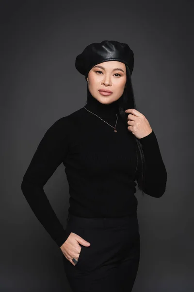 Modelo asiático de moda en boina cogida de la mano en bolsillo de pantalones aislados en gris oscuro - foto de stock