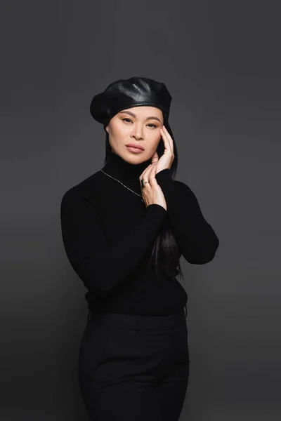 Stylish asian woman in beret and turtleneck touching face isolated on dark grey — Stock Photo