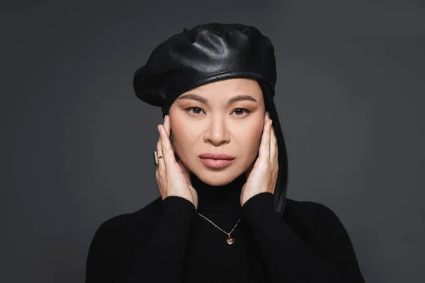 Porträt einer stilvollen asiatischen Frau mit Baskenmütze und Rollkragen, die Gesicht isoliert auf dunkelgrau berührt — Stockfoto