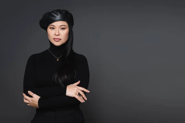 Trendy asian model in beret and necklace looking at camera isolated on dark grey — Stock Photo