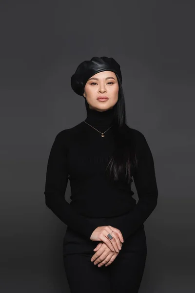 Fashionable asian woman in beret posing and looking at camera isolated on dark grey — Stock Photo