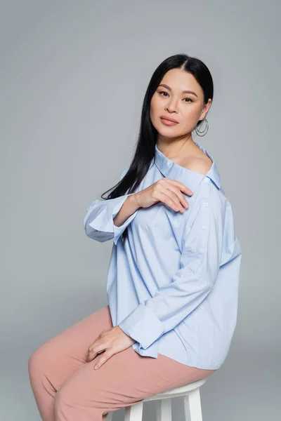 Азійка в блузці з голим плечем сидить на стільці ізольованому на сірому — стокове фото