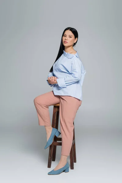 Longitud completa de elegante mujer asiática posando cerca de silla sobre fondo gris - foto de stock