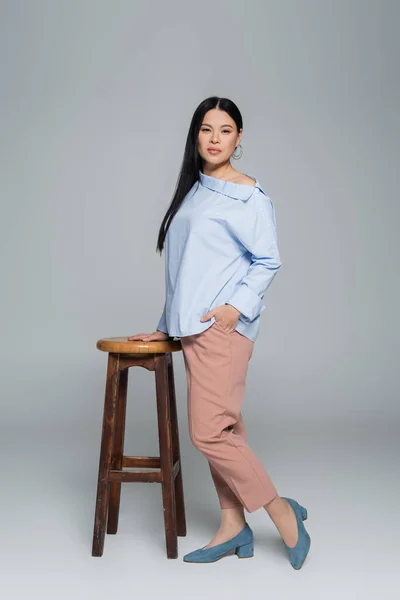 Longitud completa de mujer asiática elegante sosteniendo la mano en el bolsillo de los pantalones cerca de la silla sobre fondo gris - foto de stock