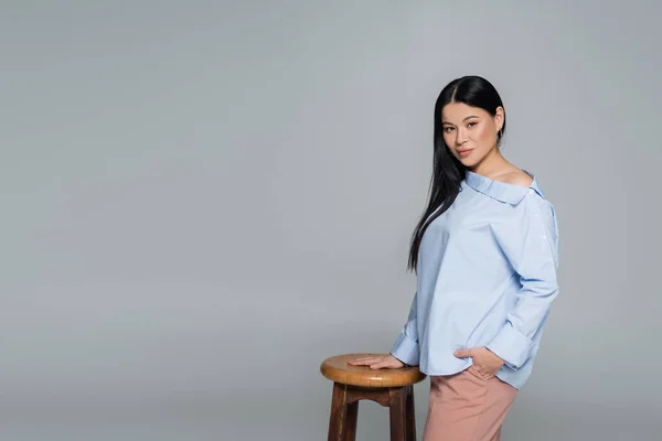 Asian model in blue blouse posing near chair isolated on grey — Stock Photo