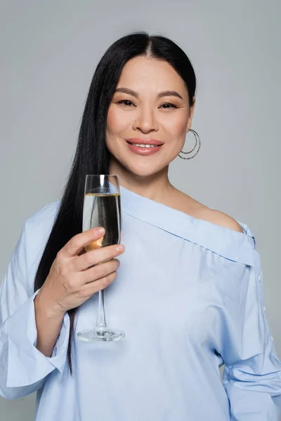 Улыбающаяся азиатка в блузке с бокалом шампанского, изолированная на сером — стоковое фото