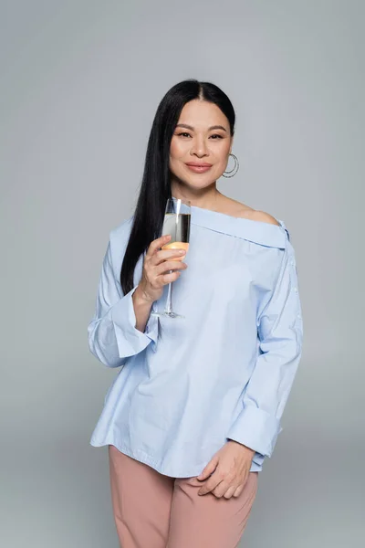 Elegante donna asiatica in possesso di un bicchiere di champagne e sorridente isolato sul grigio — Foto stock