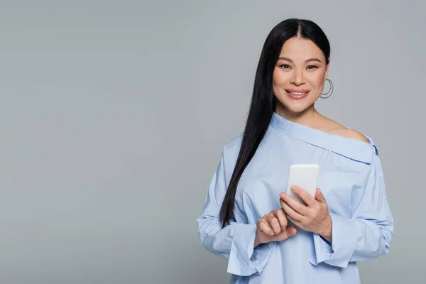 Позитивна азіатська жінка в блузці за допомогою мобільного телефону ізольована на сірому — стокове фото