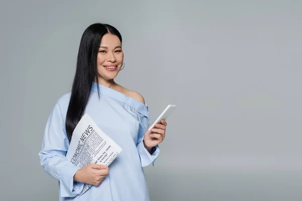 Smiling asian woman in blouse holding cellphone and economic newspaper isolated on grey — Stock Photo