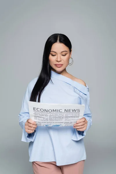 Elegante morena mujer asiática leyendo noticias económicas aislado en gris - foto de stock