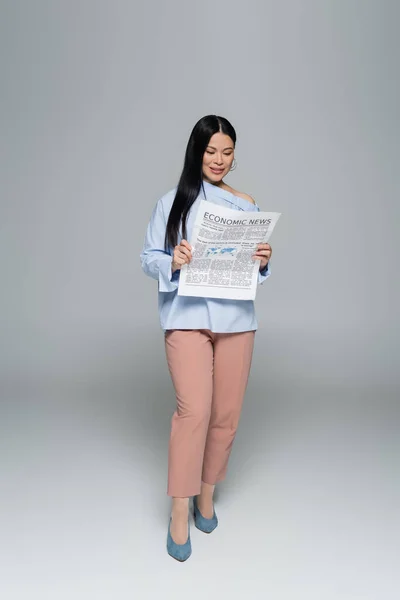 Full length of smiling asian woman reading economic newspaper on grey background — Stock Photo