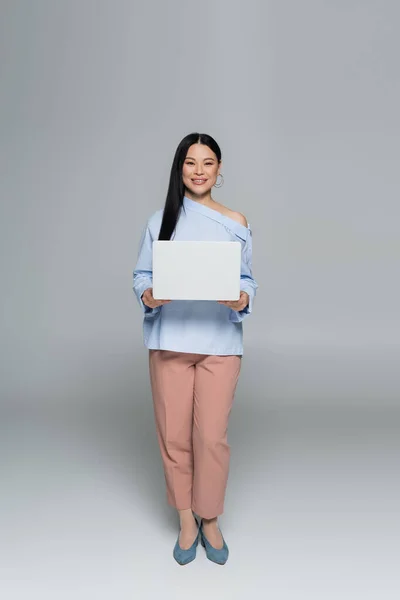 Positif et élégant asiatique femme tenant ordinateur portable sur fond gris — Photo de stock