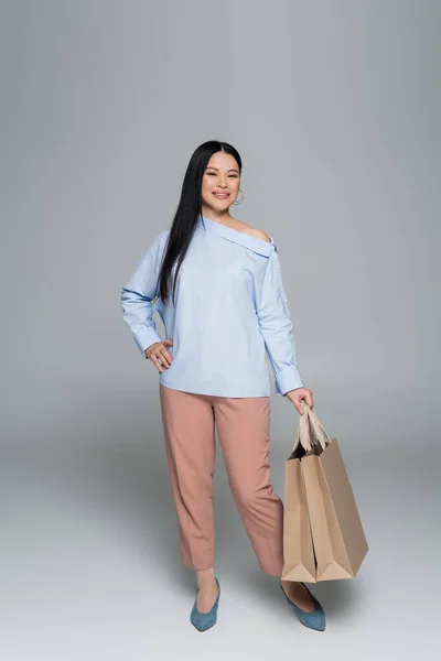 Full length of smiling asian woman holding shopping bags on grey background — Stock Photo