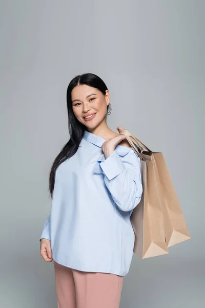Stilvolle asiatische Frau mit Einkaufstaschen und Blick in die Kamera isoliert auf grau — Stockfoto