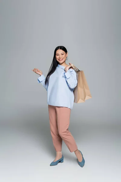Positive asian model holding shopping bags and pointing with finger on grey background — Stock Photo