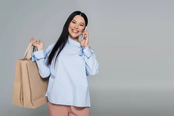 Sonriendo mujer asiática sosteniendo bolsas de compras y hablando en el teléfono inteligente aislado en gris - foto de stock
