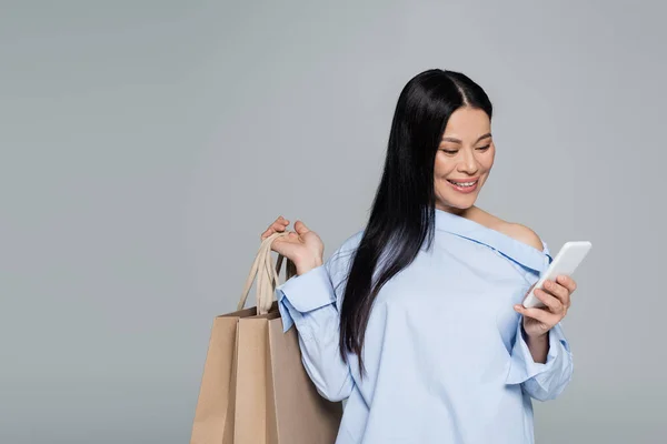 Positiva donna asiatica utilizzando smartphone e tenendo borse della spesa isolate sul grigio — Foto stock