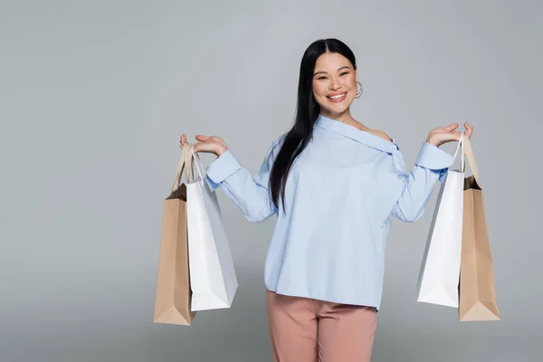 Cheerful asian woman in blouse holding purchases isolated on grey — Stock Photo
