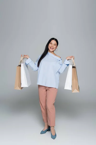 Longitud completa de sonriente positiva mujer asiática sosteniendo bolsas de compras sobre fondo gris - foto de stock